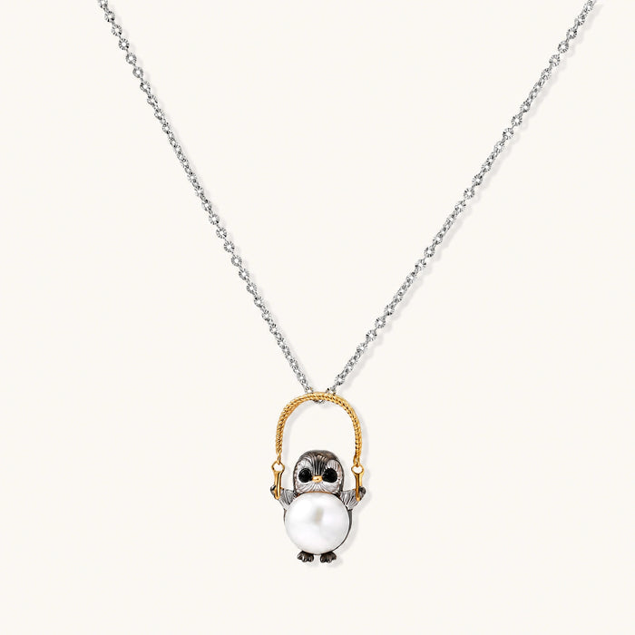 Whimsical penguin pendant holding a white pearl on a gold swing, rhodium-plated silver chain on cream background.