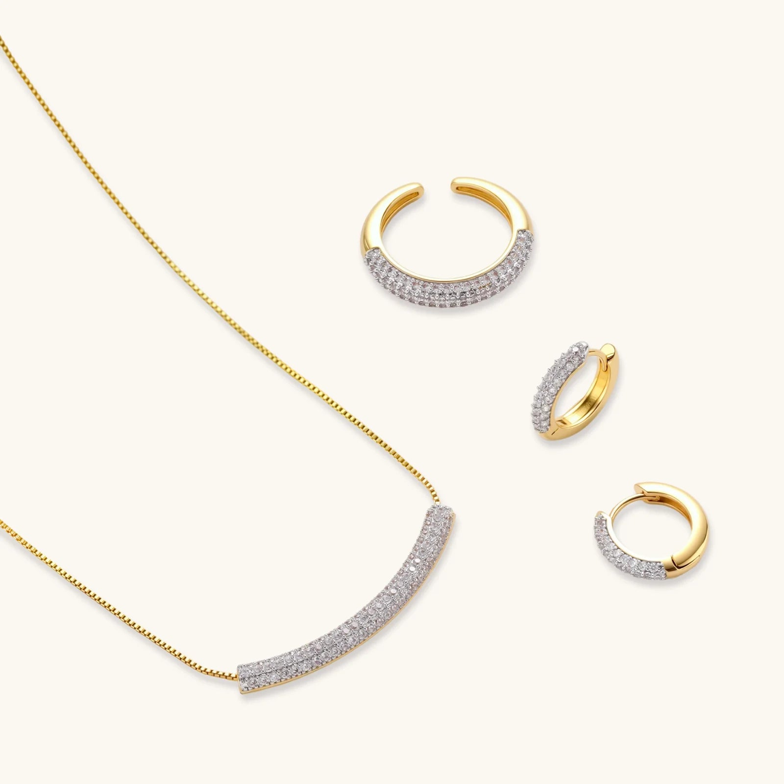 Gold-plated curved necklace, hoop earrings, and adjustable ring with pavé cubic zirconia stones on cream background.