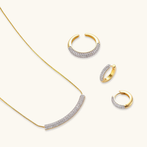 Gold-plated curved necklace, hoop earrings, and adjustable ring with pavé cubic zirconia stones on cream background.