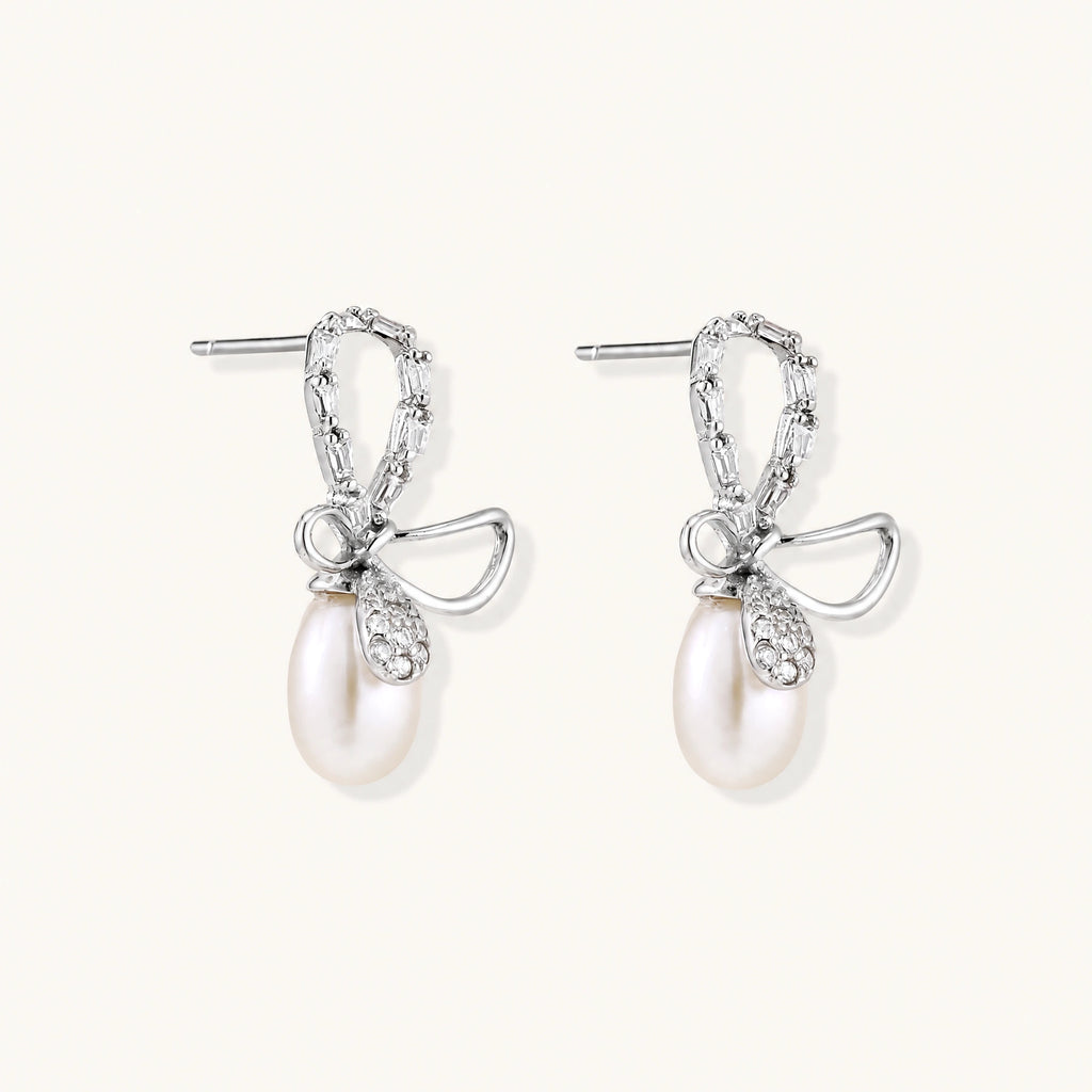 Silver bow-shaped stud earrings with baguette crystals and dangling white freshwater pearls on cream background
