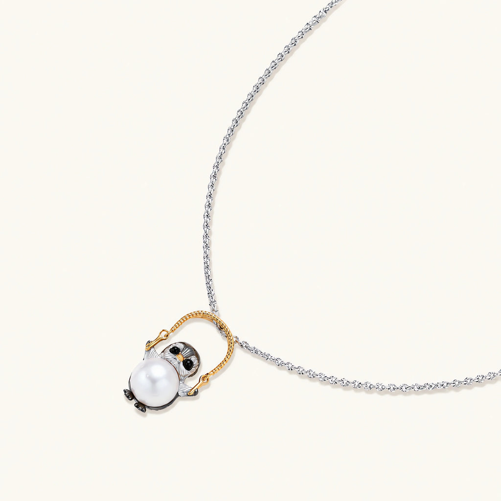 Whimsical penguin pendant holding a white pearl on a gold swing, rhodium-plated silver chain on cream background.