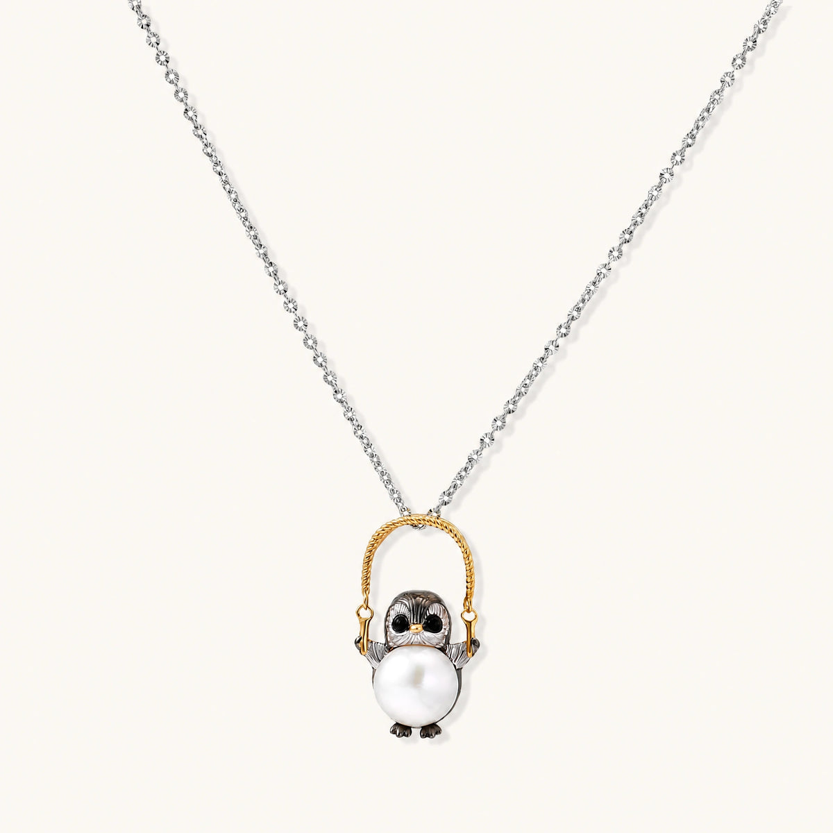 Whimsical penguin pendant holding a white pearl on a gold swing, rhodium-plated silver chain on cream background.