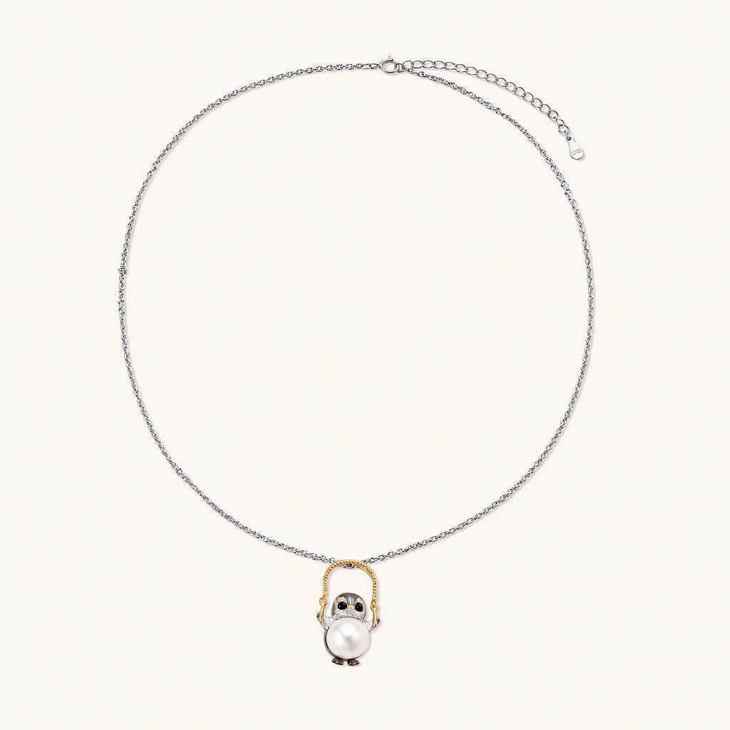 Whimsical penguin pendant holding a white pearl on a gold swing, rhodium-plated silver chain on cream background.